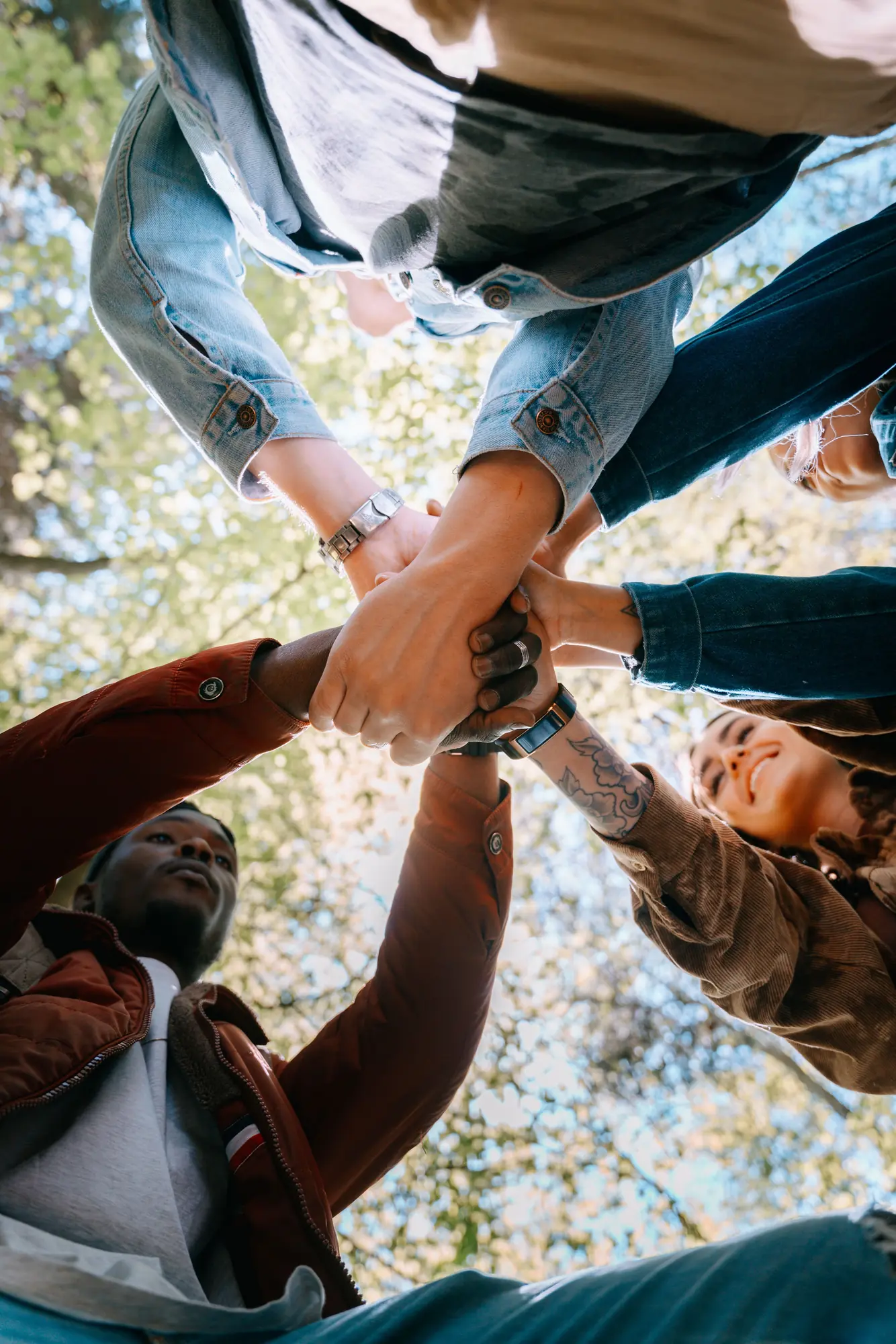 A group photographed from below under trees, joining their hands above the camera.